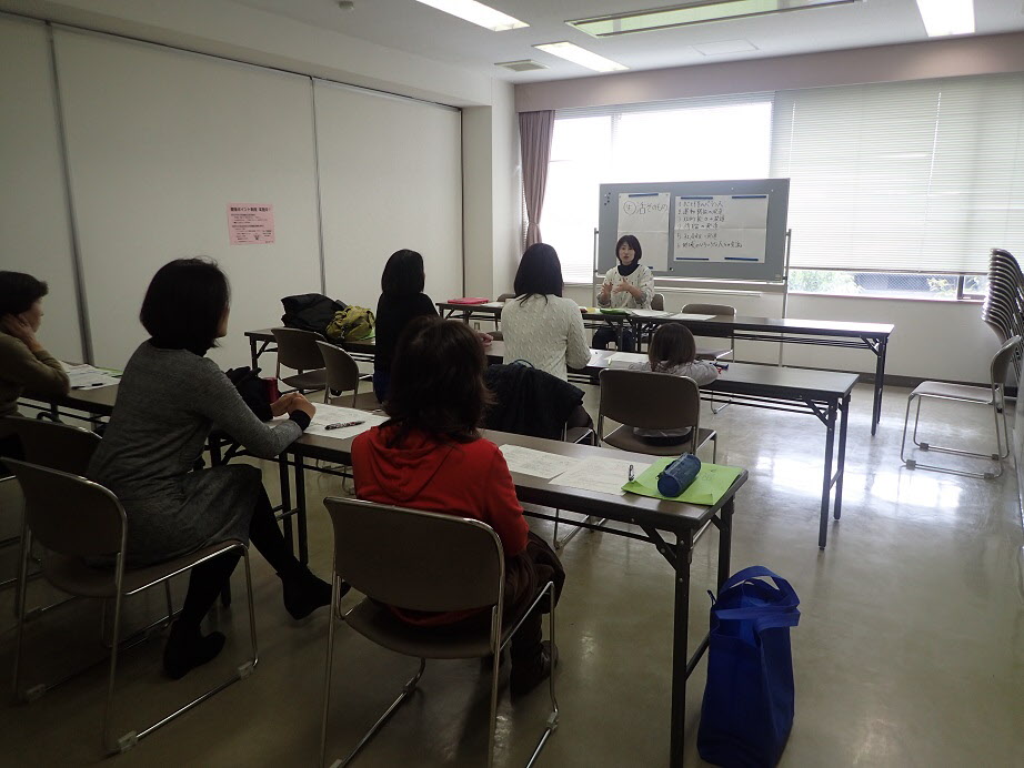 ボードの前で説明をしている女性の話を長机に座った支援会員の5名が聞いている講習会の様子を部屋の後方から写した写真