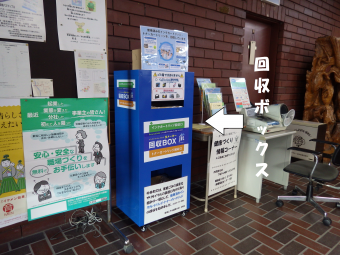 役場の玄関にあるチラシが貼ってあるボードの隣の青いインクカートリッジ回収箱の写真