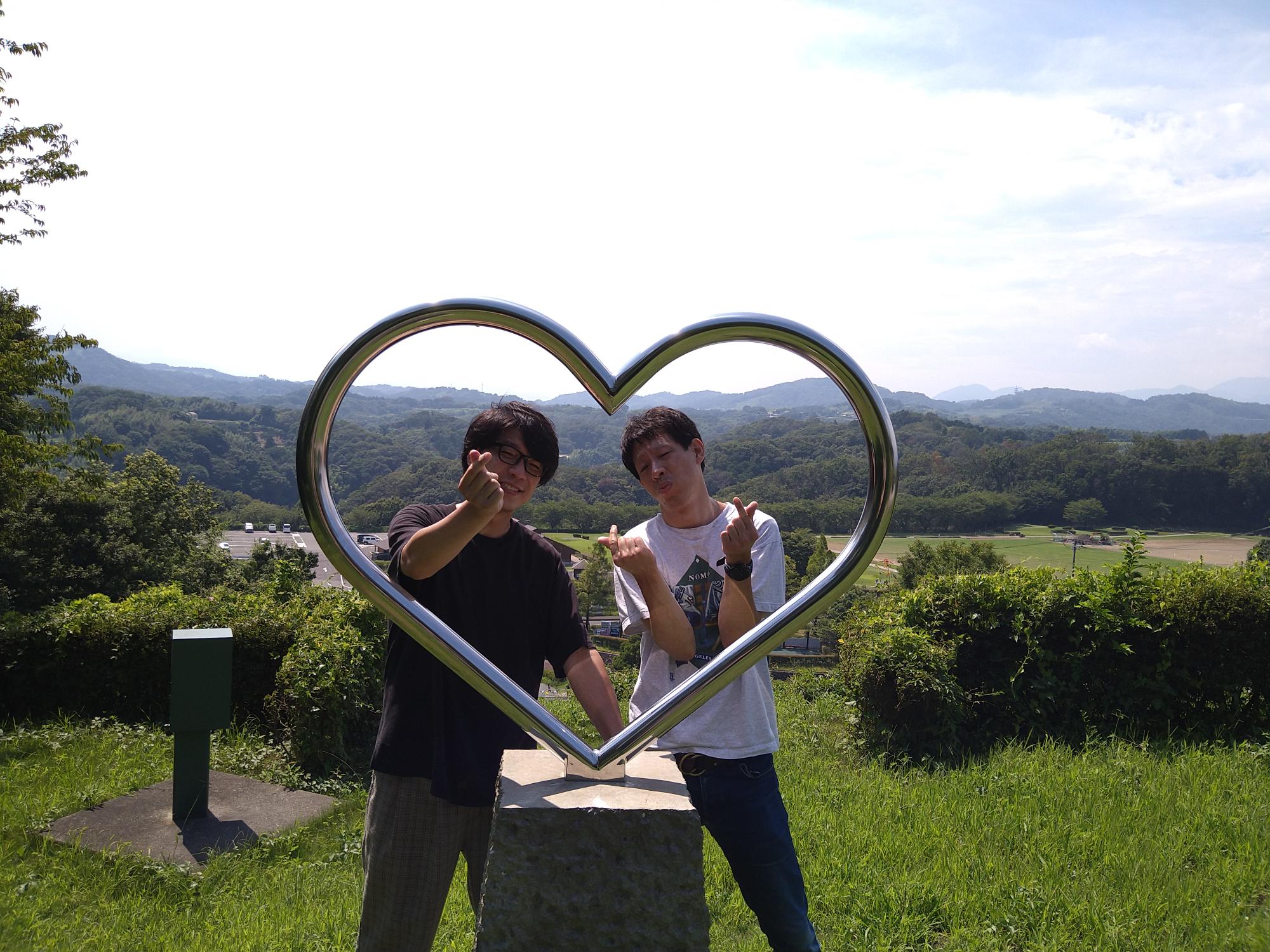 芸人囲碁将棋の二人が中井中央公園内のハートの丘にてハートのオブジェ越しに記念撮影をしている